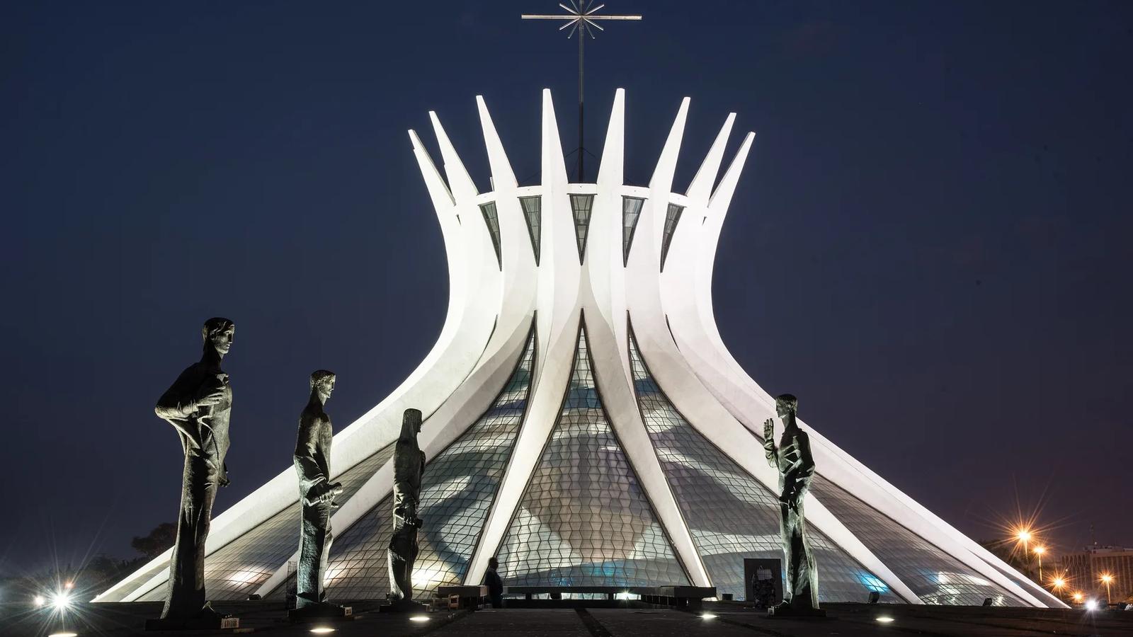 Catedral de Brasília iluminada à noite, com as colunas curvas de concreto e quatro estátuas de bronze dos evangelistas na praça em primeiro plano