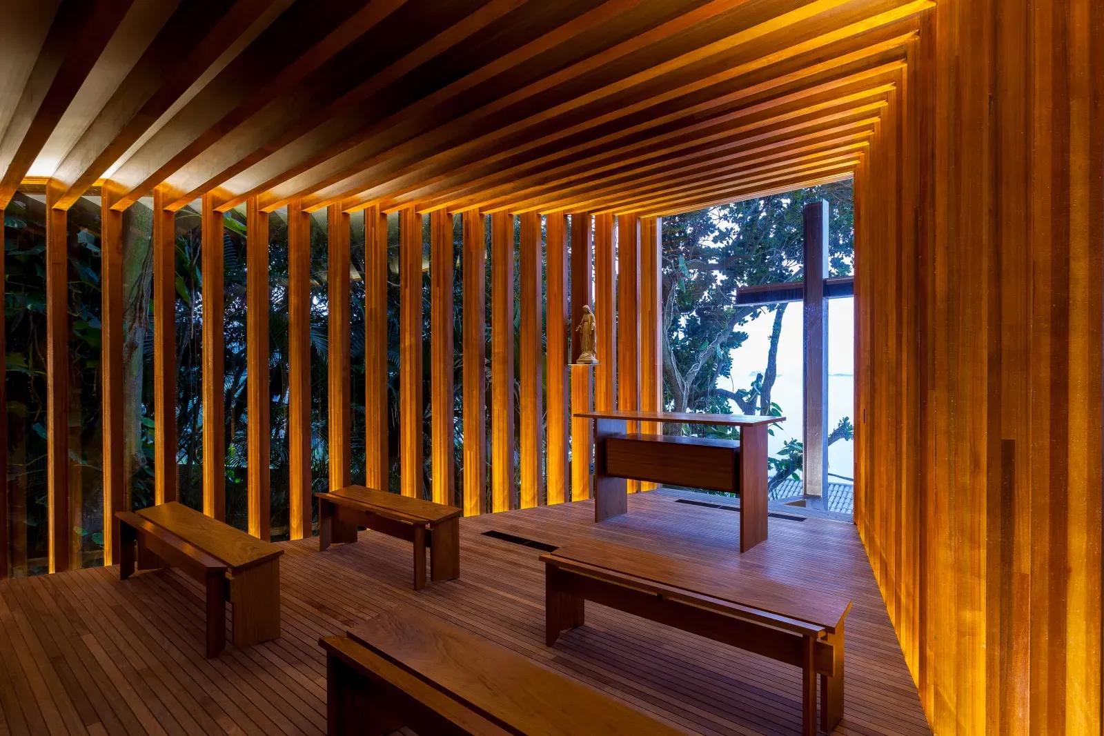 Interior de pequena capela revestida de ripas de madeira, com bancos de madeira e vista para a mata