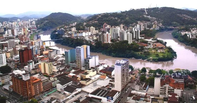 Vista aérea do Centro de Blumenau com prédios residenciais, uma curva do rio Itajaí-Açu e morros cobertos de mata ao fundo