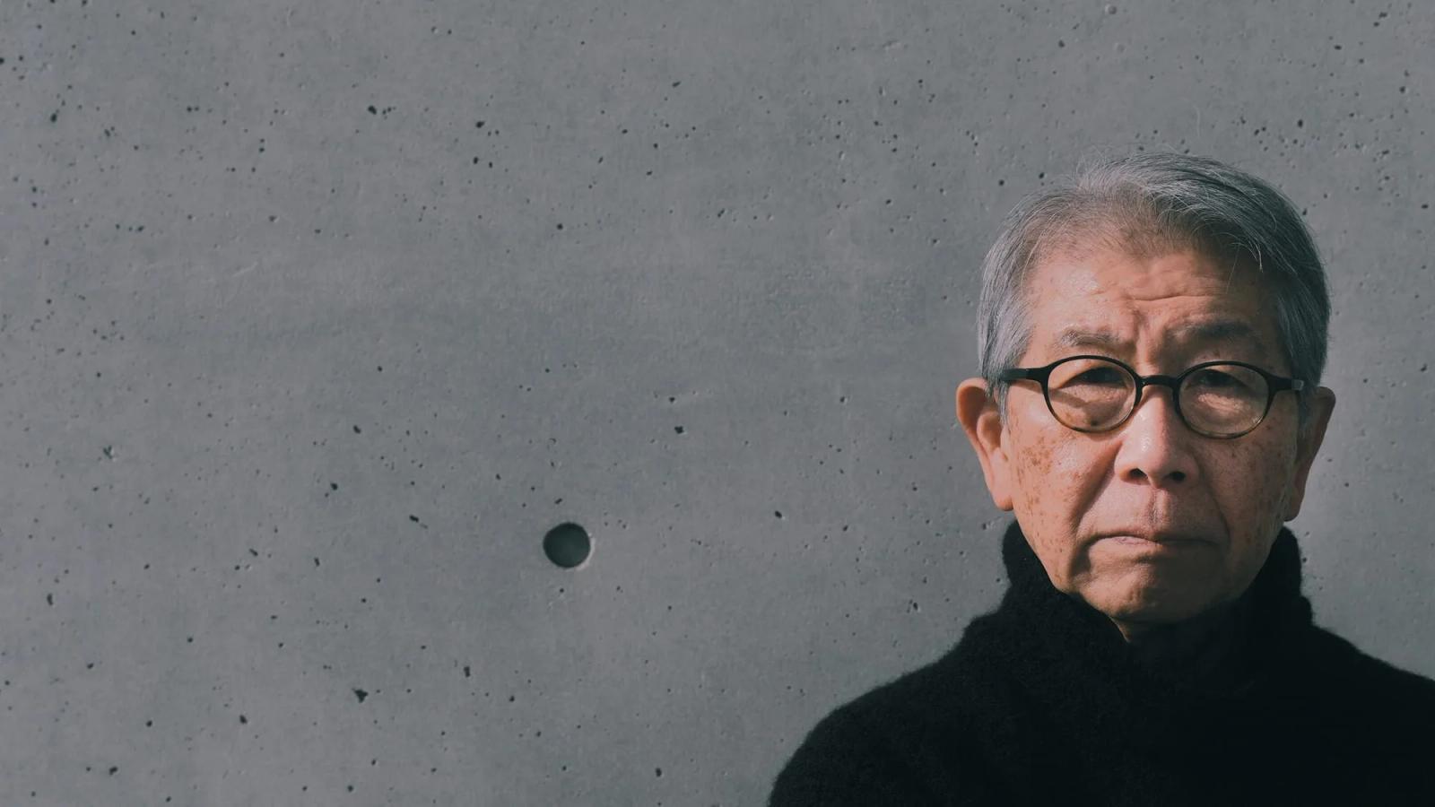 Retrato do arquiteto Riken Yamamoto, de cabelos brancos, óculos redondos e gola rulê preta, diante de uma parede de concreto aparente.