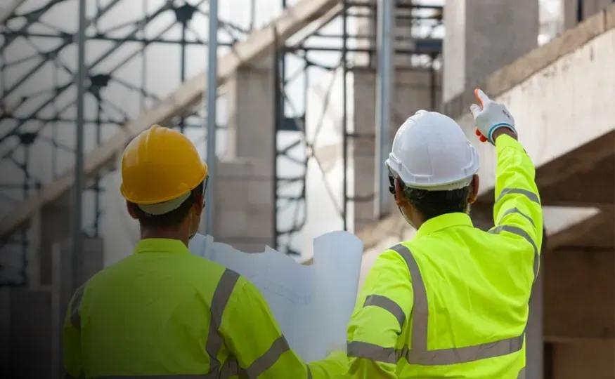 Dois profissionais de capacete e colete refletivo, de costas, observando a estrutura de um prédio em construção