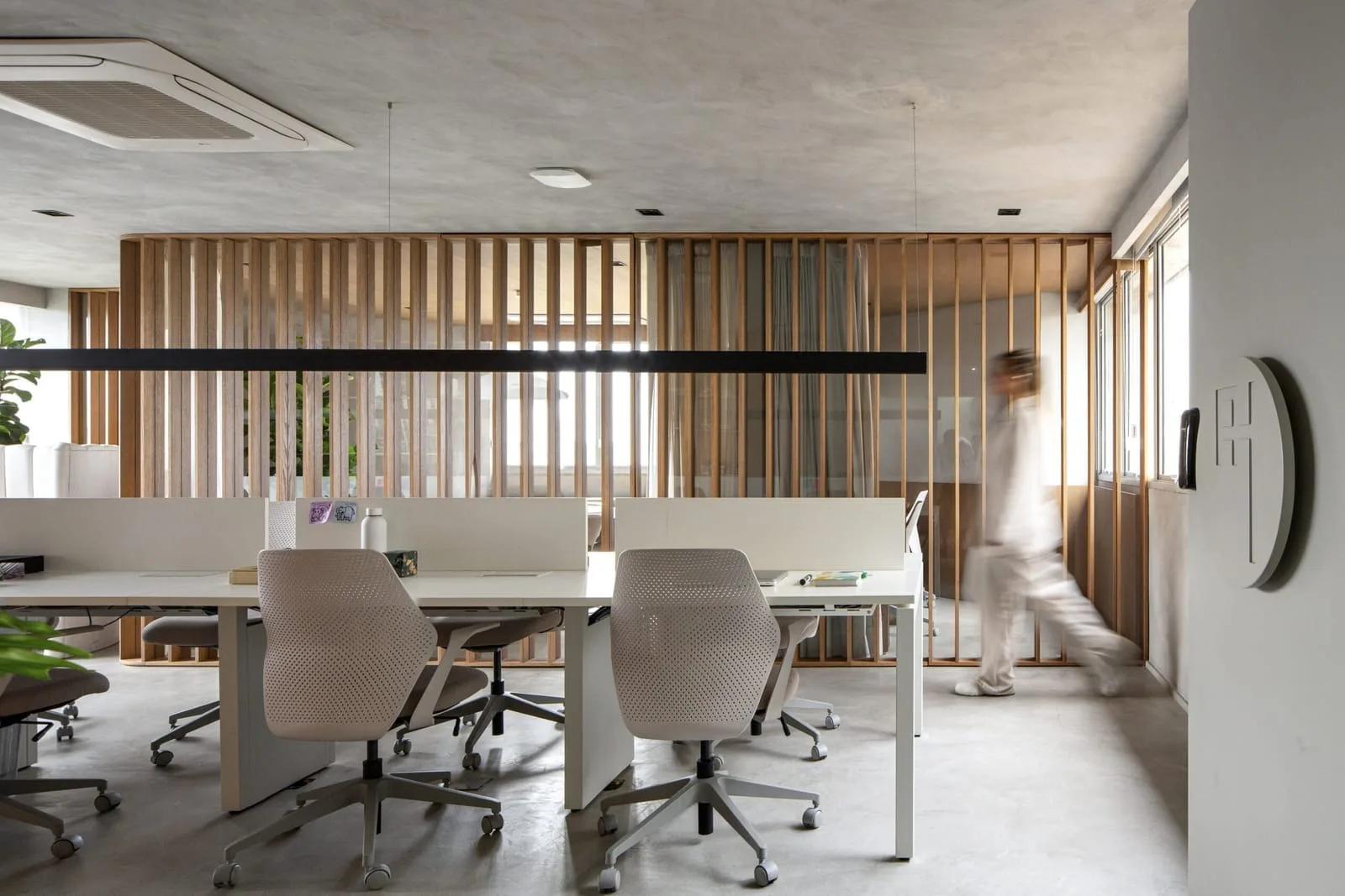 Escritório contemporâneo Escritório com piso de concreto, mesas brancas com cadeiras cinza, divisória de ripas de madeira e uma pessoa passando em movimento ao fundo