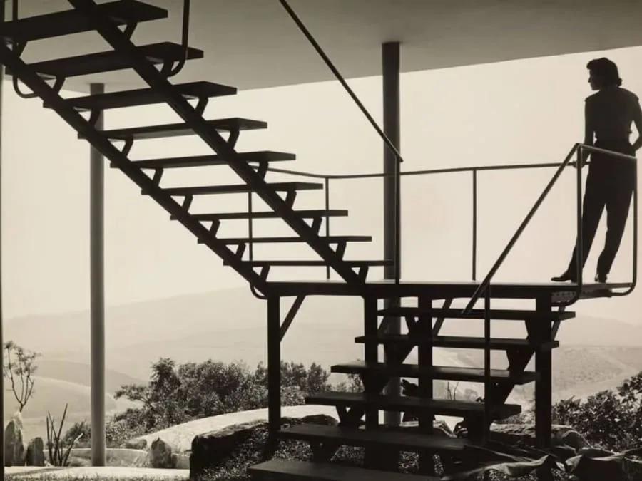 Lina na Casa de Vidro Lina Bo Bardi de pé no alto da escada metálica da Casa de Vidro, em foto em preto e branco
