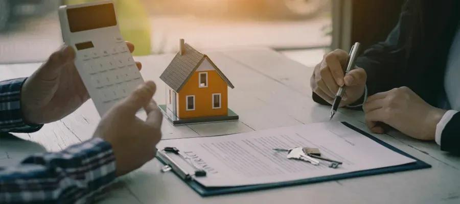 Contratação de financiamento com alienação fiduciária Duas pessoas à mesa com calculadora, casa em miniatura, chaves e contrato para assinatura