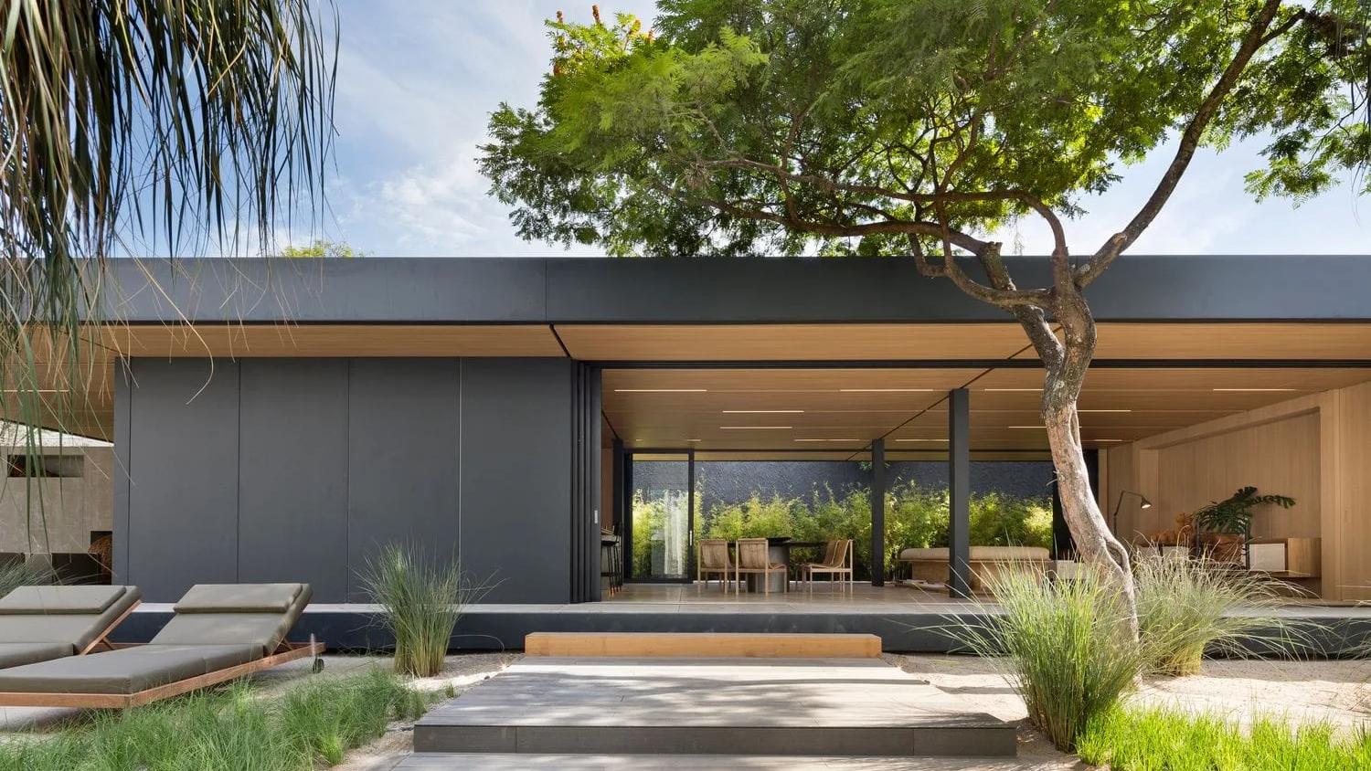 Casa-modelo SysHaus, do Studio Arthur Casas, em São Paulo Casa SysHaus de fachada escura e cobertura plana, com a sala aberta para o jardim de capim e uma árvore em primeiro plano