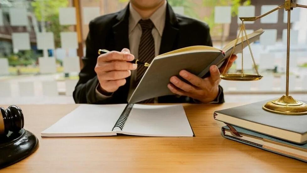 Profissional de terno escreve em um caderno sobre a mesa, ao lado de livros, uma balança da justiça e um martelo de juiz
