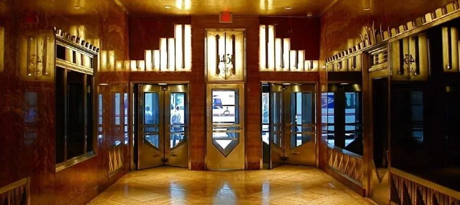 Saguão com paredes de mármore avermelhado, portas de elevador decoradas e luminárias geométricas