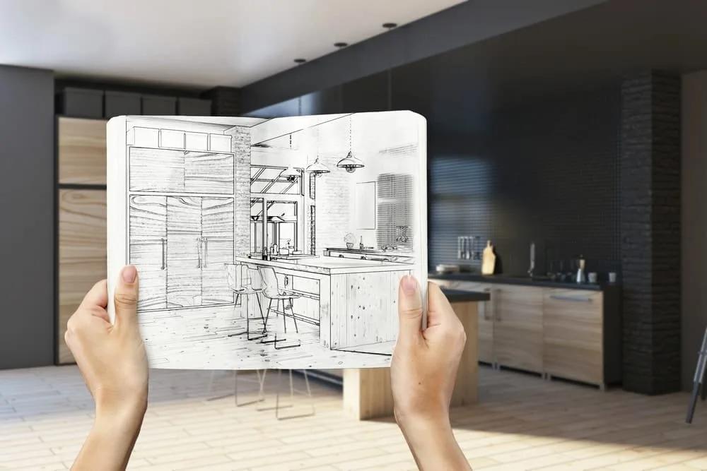 Mãos segurando um caderno com o croqui de uma cozinha diante da mesma cozinha já pronta ao fundo