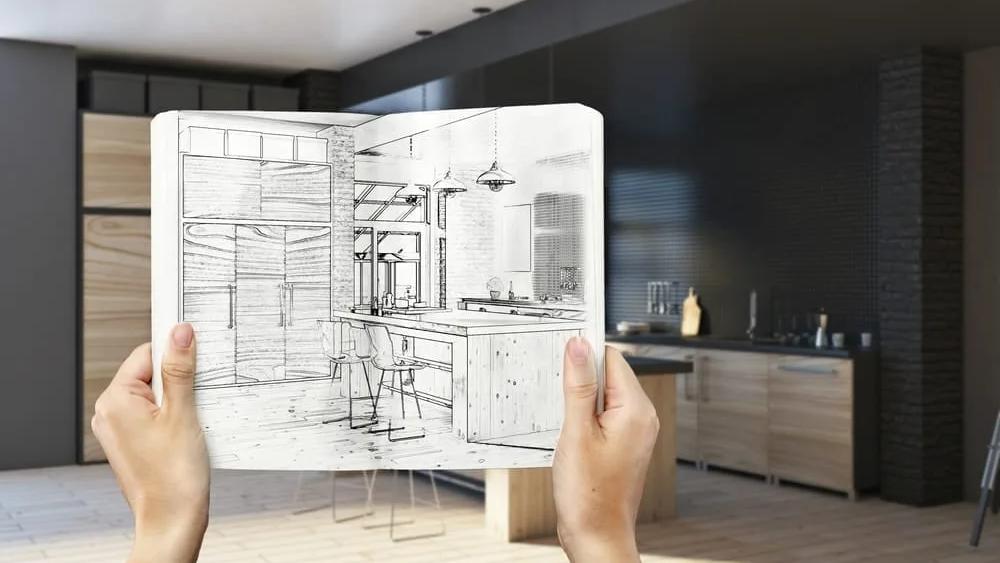 Mãos segurando um caderno com o croqui de uma cozinha diante da mesma cozinha já pronta ao fundo