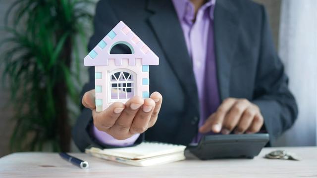 Homem de terno segura na palma da mão uma miniatura de casa; sobre a mesa, uma calculadora e um caderno.