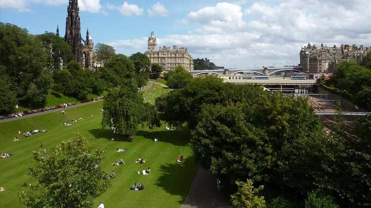Jardins da Princes Street com gramado ocupado por visitantes, árvores e, ao fundo, o Hotel Balmoral e o Scott Monument