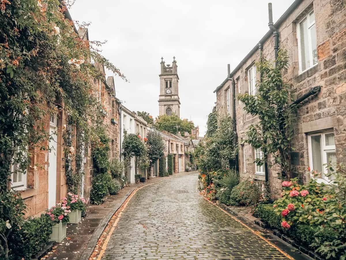 Circus Lane, rua estreita de paralelepípedos com casas baixas de pedra cobertas de trepadeiras e flores, e uma torre de igreja ao fundo