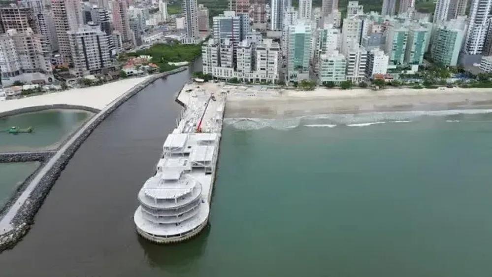 Vista aérea da orla de Itapema com um píer em obras avançando sobre o mar, prédios altos na orla e a foz de um rio protegida por molhe