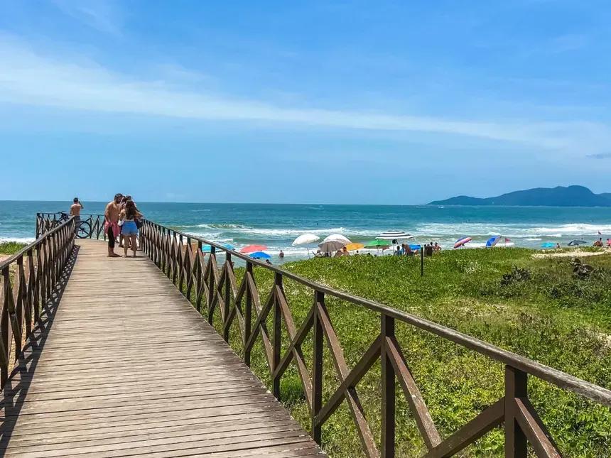 Passarela de acesso à praia Passarela de madeira sobre a restinga com banhistas caminhando em direção a uma praia movimentada, com morros ao fundo