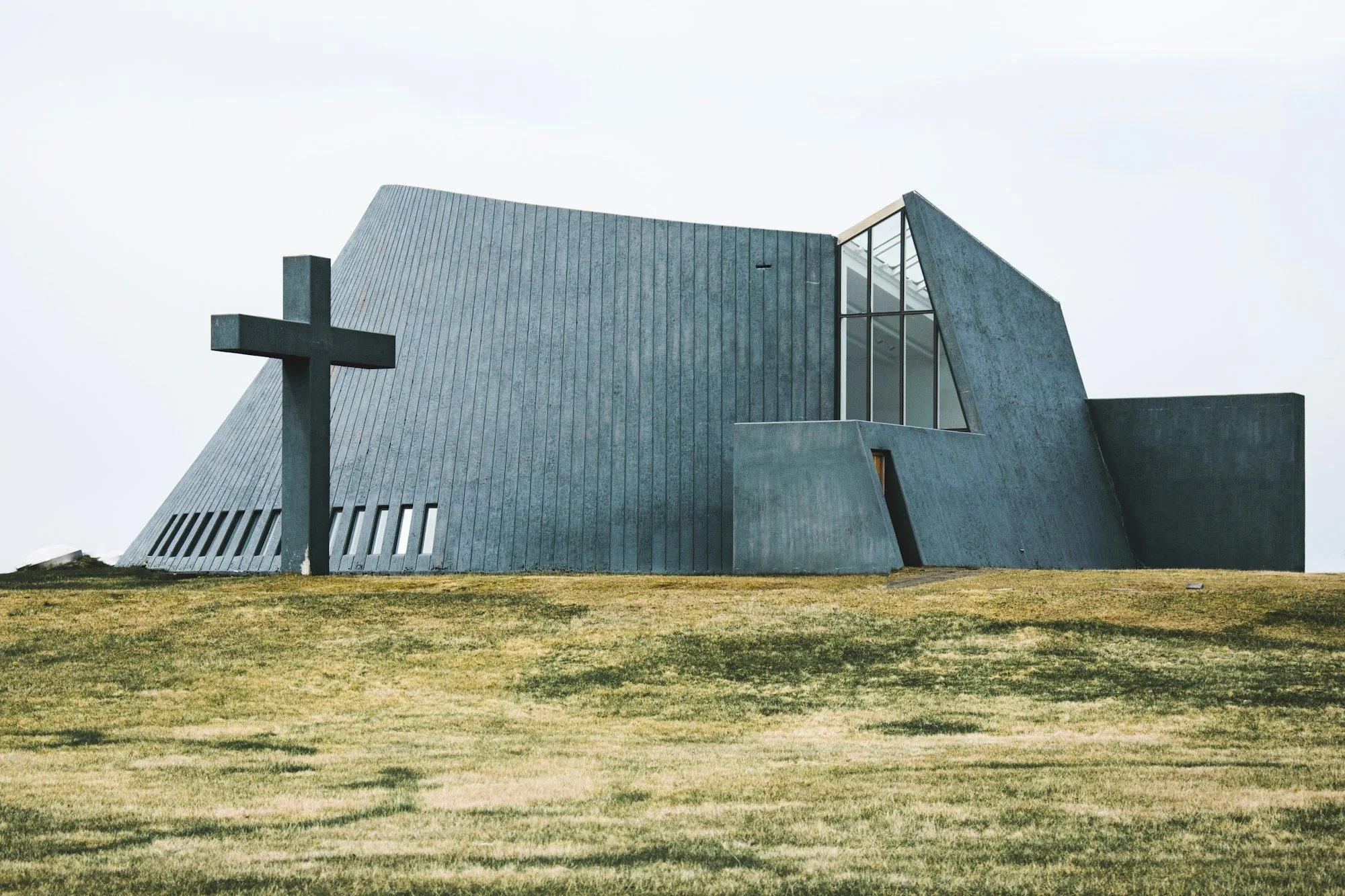 Igreja de concreto cinza com volumes inclinados e uma grande cruz ao lado, sobre um campo gramado