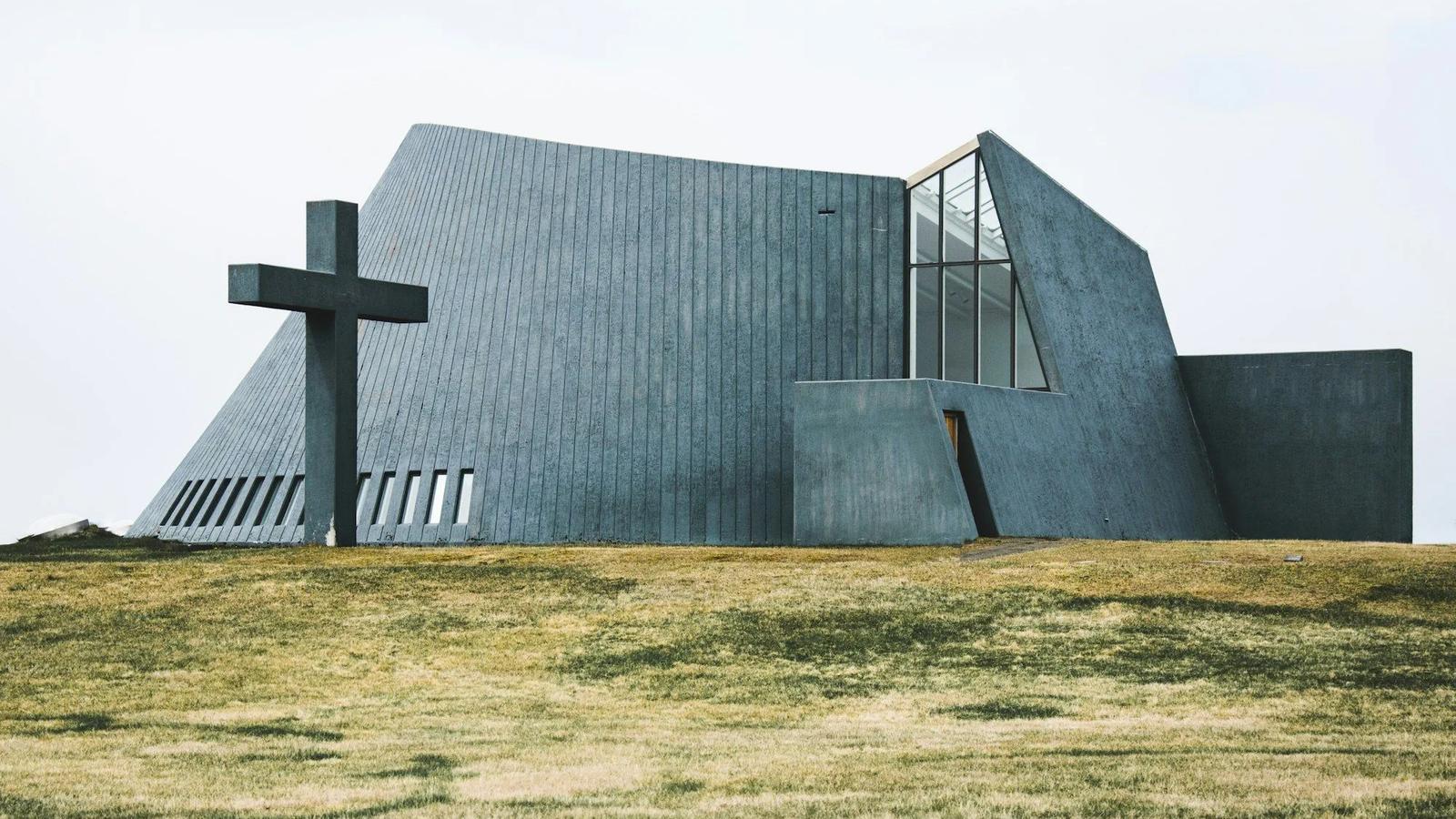 Igreja de concreto cinza com volumes inclinados e uma grande cruz ao lado, sobre um campo gramado