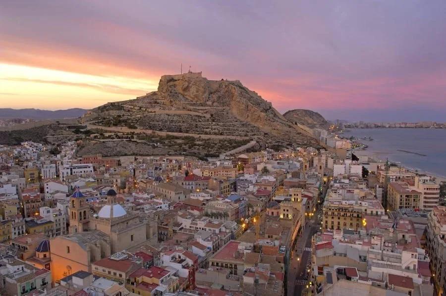 Centro antigo de Alicante com casas baixas e cúpulas de igreja ao pé de um morro rochoso com um castelo no alto, ao entardecer, junto ao mar
