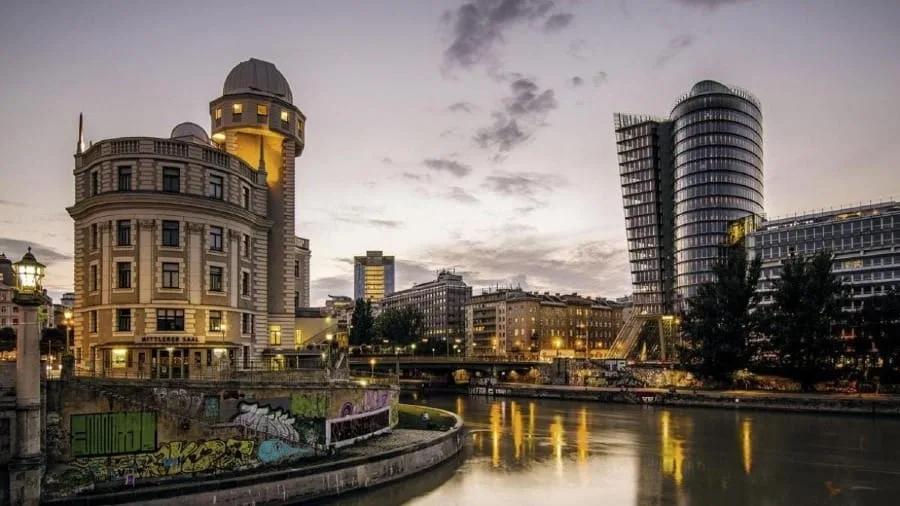 Observatório Urania, edifício Art Nouveau com torre e cúpula às margens do canal do Danúbio, com torres envidraçadas modernas na outra margem ao entardecer
