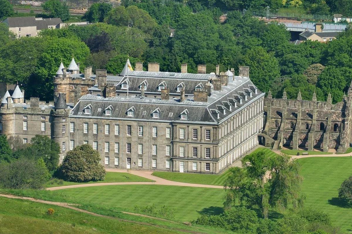 Palácio de Holyroodhouse visto do alto, com a fachada de pedra, as torres de canto e as ruínas da abadia ao lado, cercado por gramados