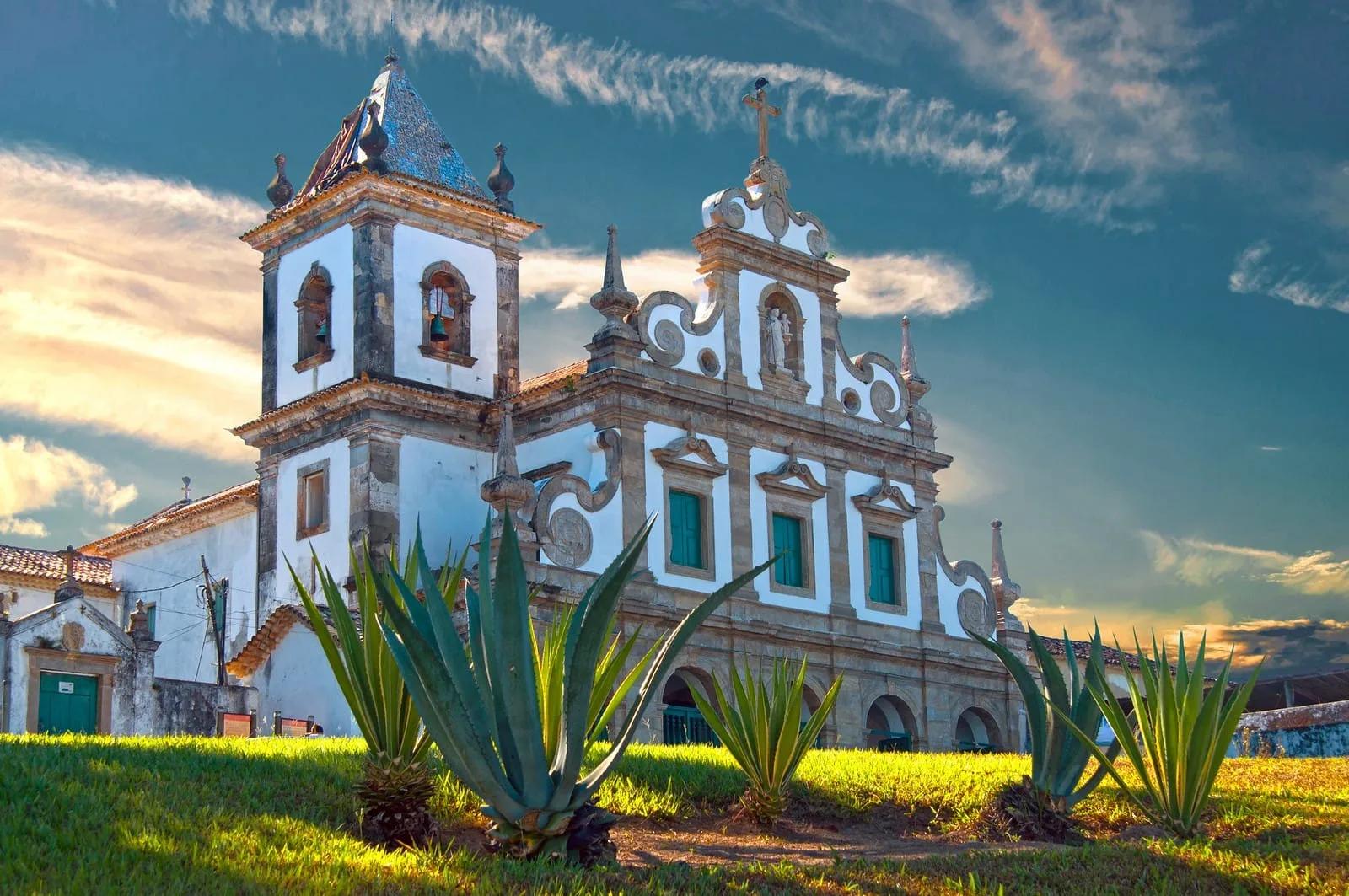Igreja barroca no Brasil Igreja colonial branca com ornamentos azuis e torre sineira, cercada de agaves