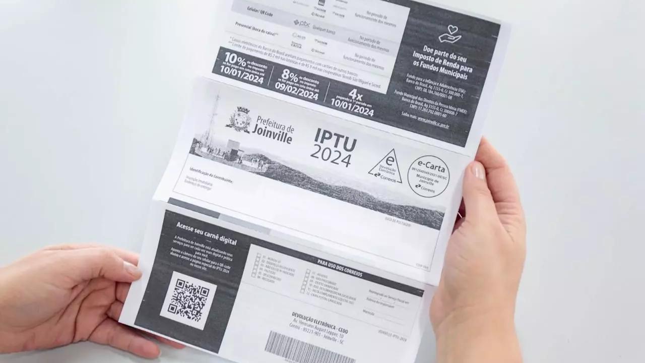 Mãos segurando o carnê do IPTU 2024 da Prefeitura de Joinville, com QR Code e opções de pagamento