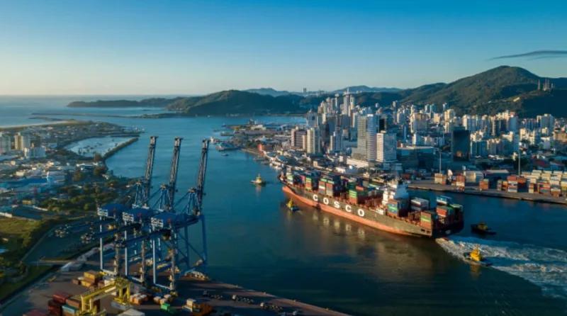 Navio porta-contêineres entrando pelo rio Itajaí-Açu, com portêineres na margem de Navegantes e a cidade de Itajaí ao fundo