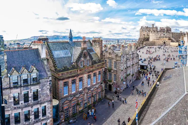 Royal Mile Trecho alto da Royal Mile com fachadas de pedra e telhados de ardósia, visto do alto, com a esplanada do castelo ao fundo e pedestres na rua