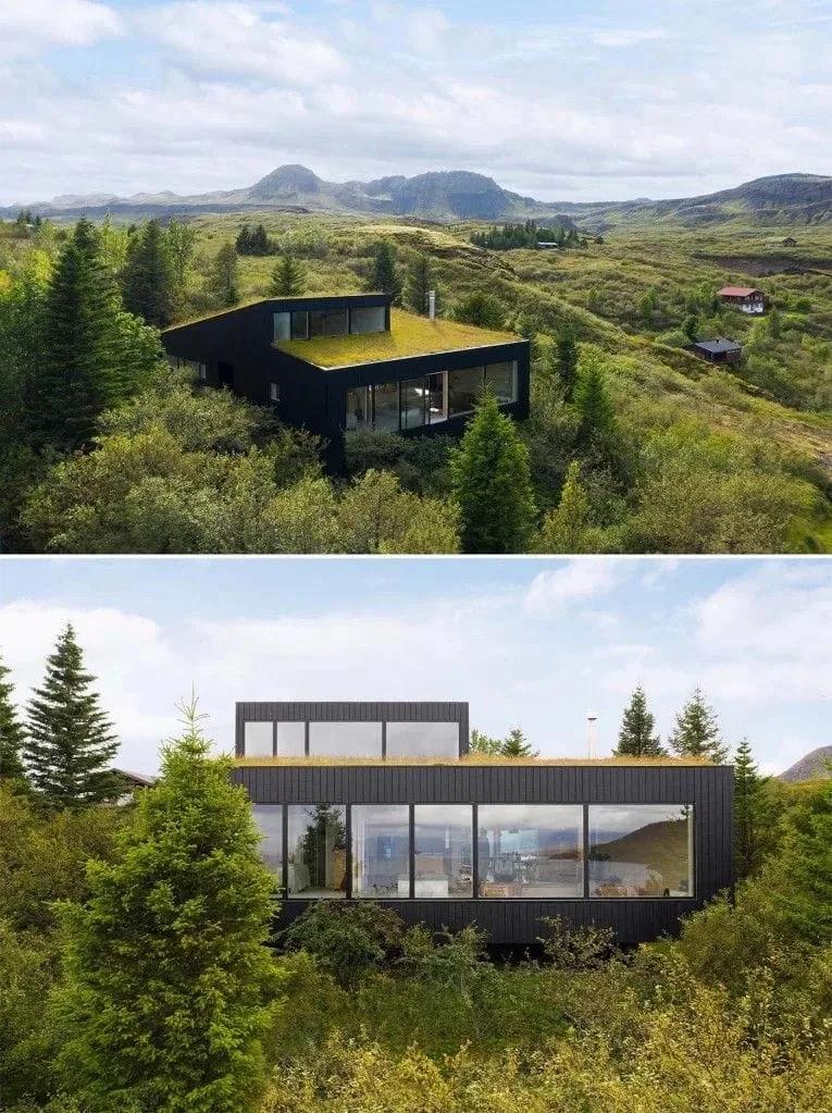 Casa de volumes escuros com telhado verde e grandes janelas, inserida em paisagem de colinas e mata