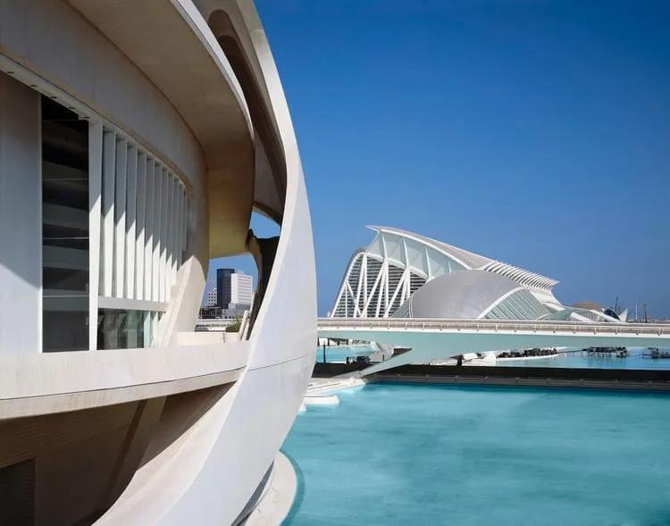Detalhe curvo de um edifício branco em primeiro plano e o Museu de les Ciències ao fundo, com espelho d'água azul