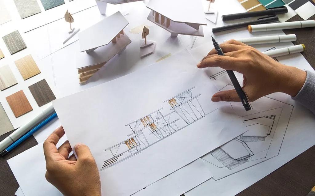 Estudo de projeto com desenho, maquete e amostras de materiais Mãos desenhando um projeto à caneta sobre uma prancheta, com maquetes e amostras de materiais ao redor