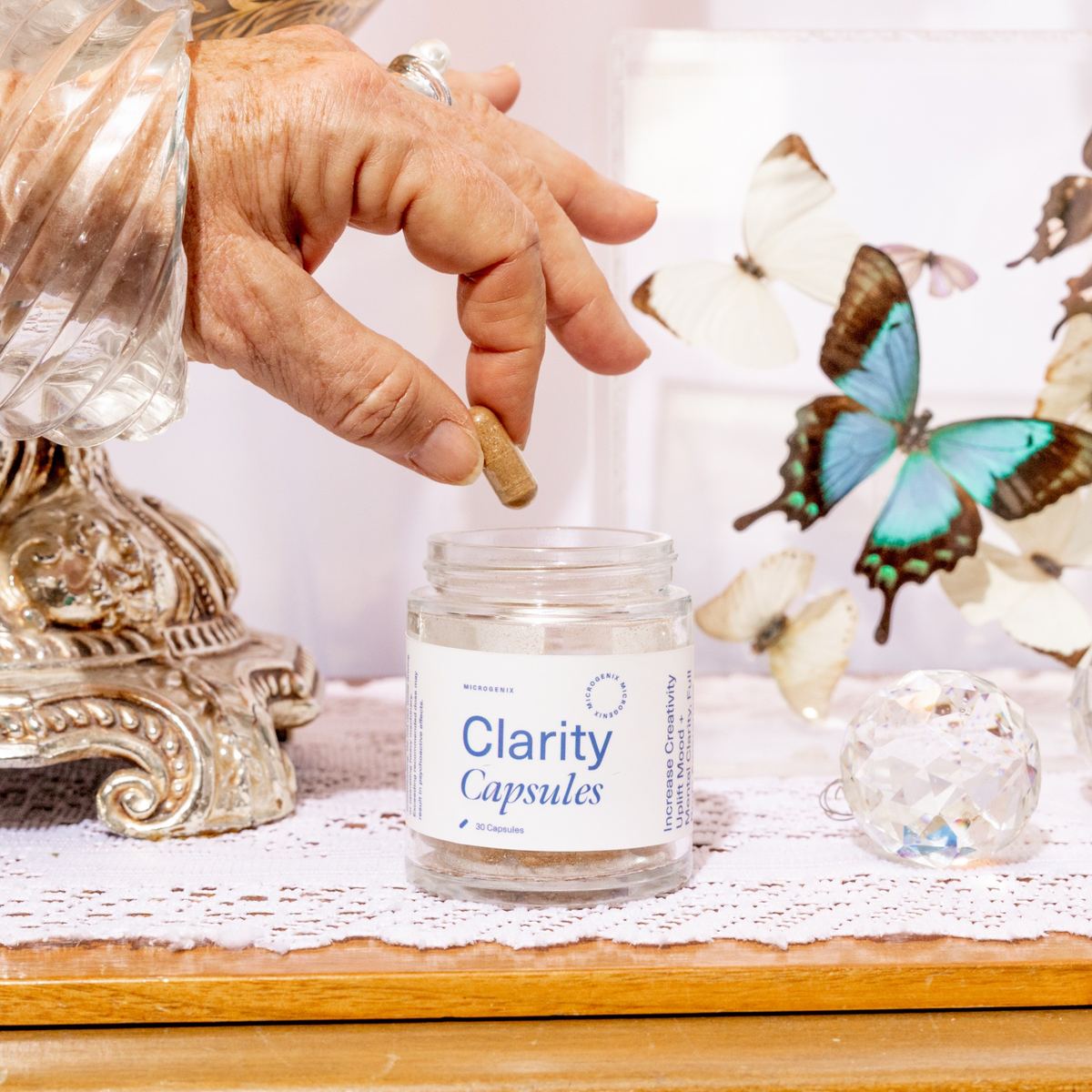 Hand lifting a capsule from a Clarity Capsules jar surrounded by butterflies