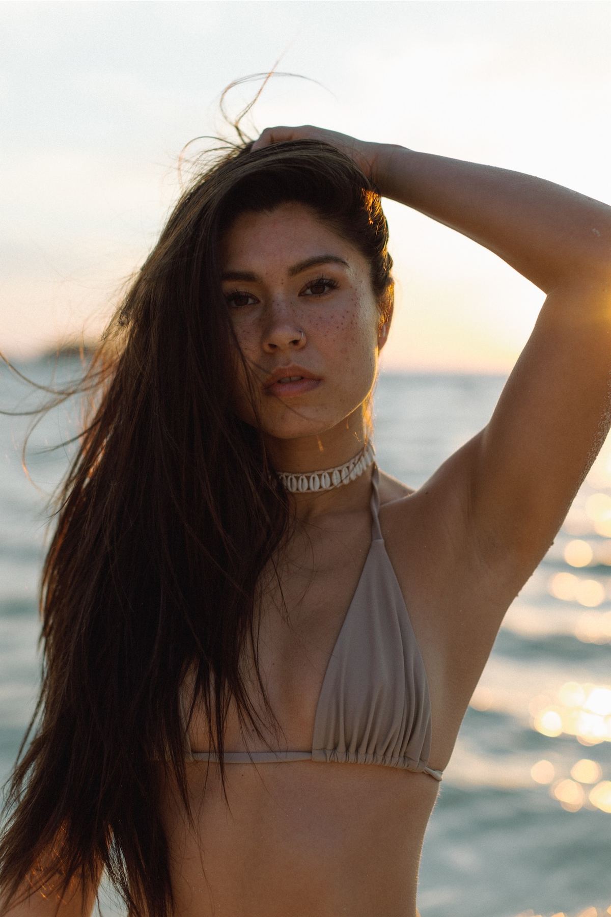 Model in a pale bikini running a hand through her hair with the sea behind her
