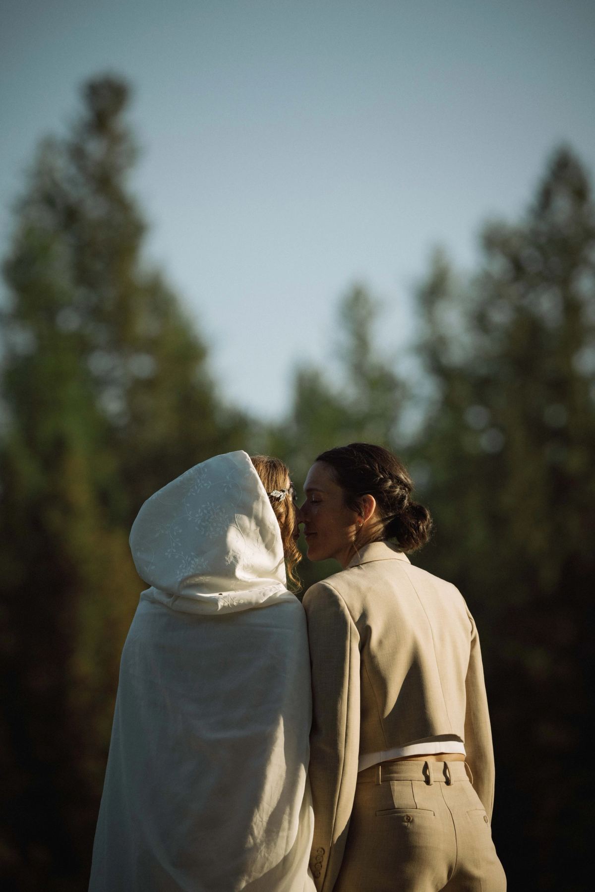 Two brides facing each other among the pines, one in a hooded veil