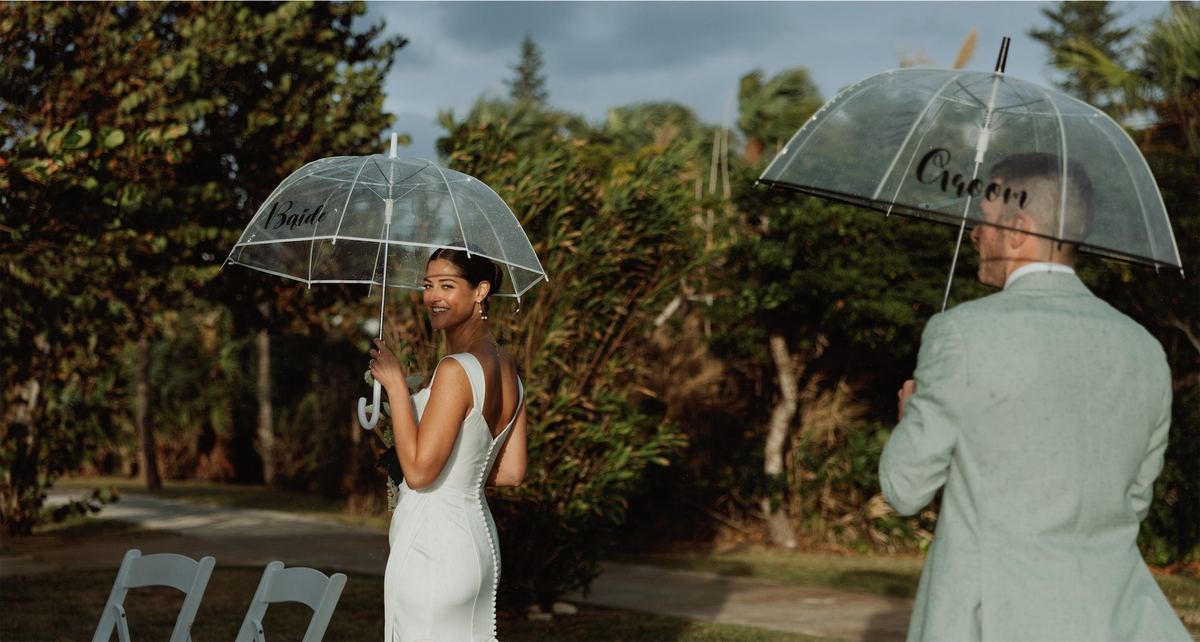 Bride and groom sheltering under clear umbrellas on the lawn in the rain