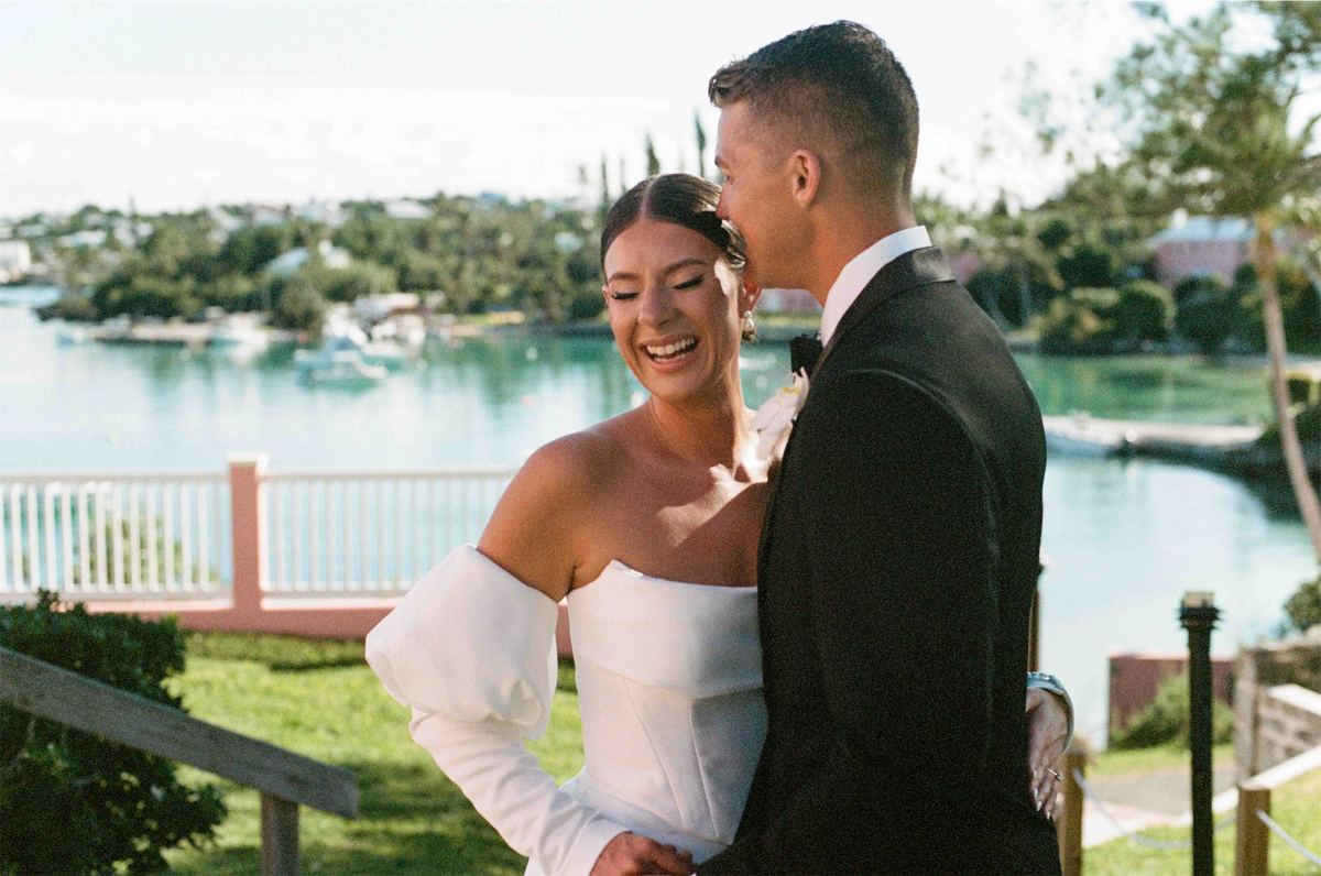 Bride laughing as her groom kisses her cheek on the lawn above the harbour