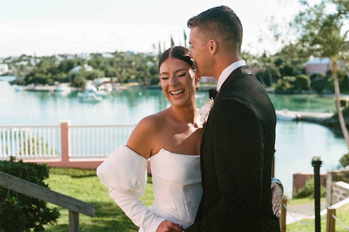 Bride laughing as her groom kisses her cheek on the lawn above the harbour