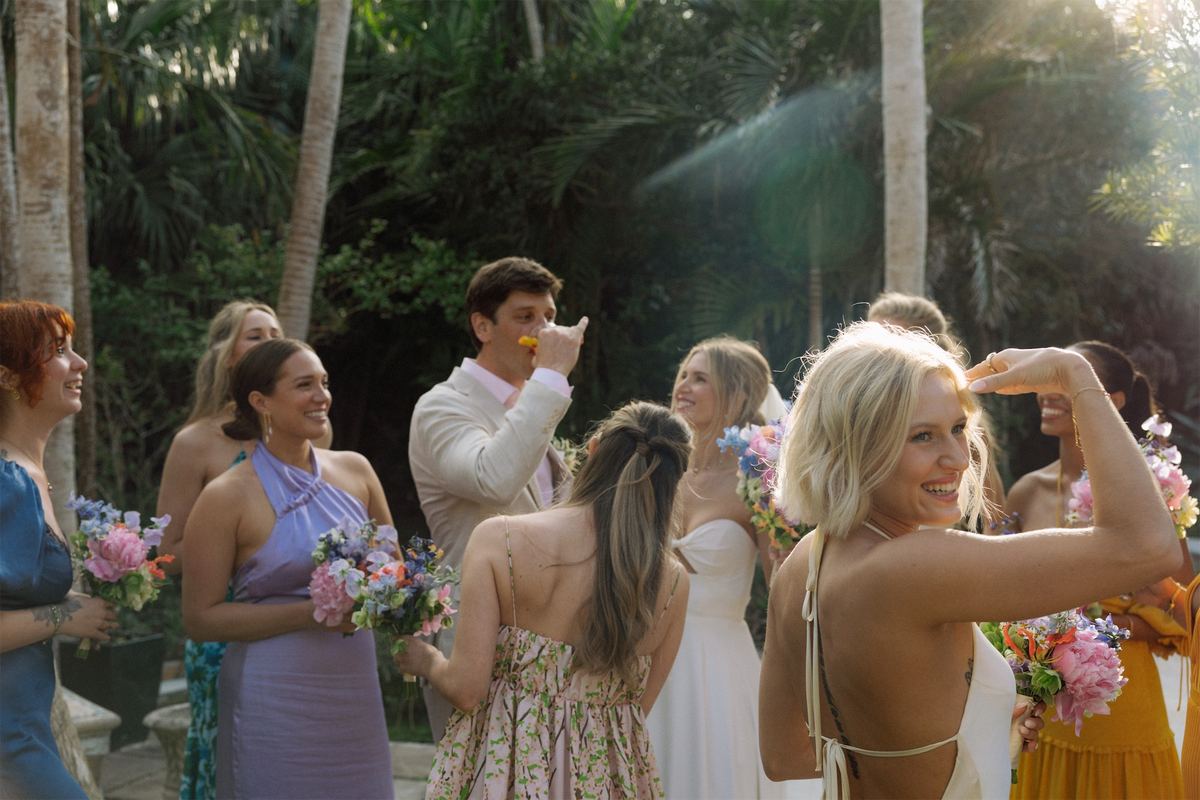 Groom toasting with bridesmaids holding pastel bouquets in a palm-shaded garden