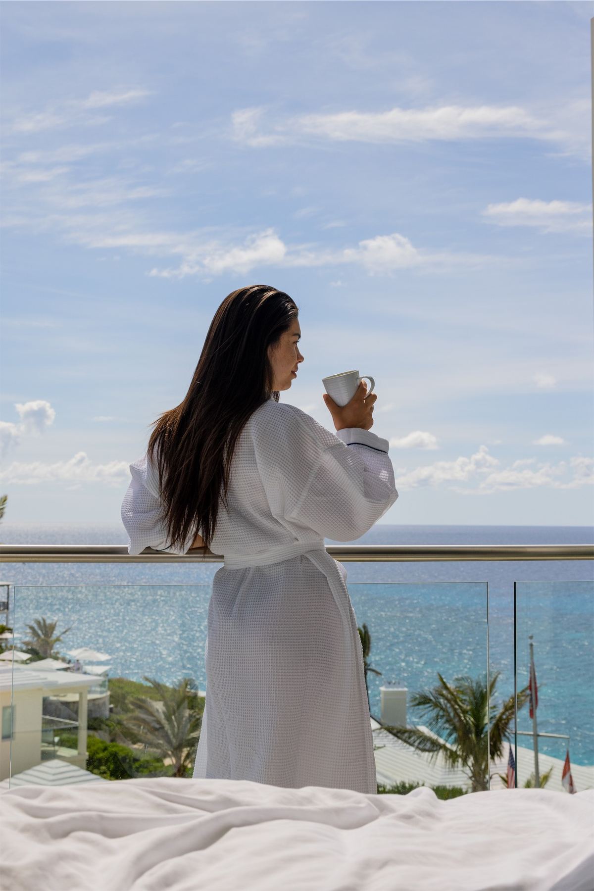 Guest in a white robe holding coffee on a balcony facing the open ocean