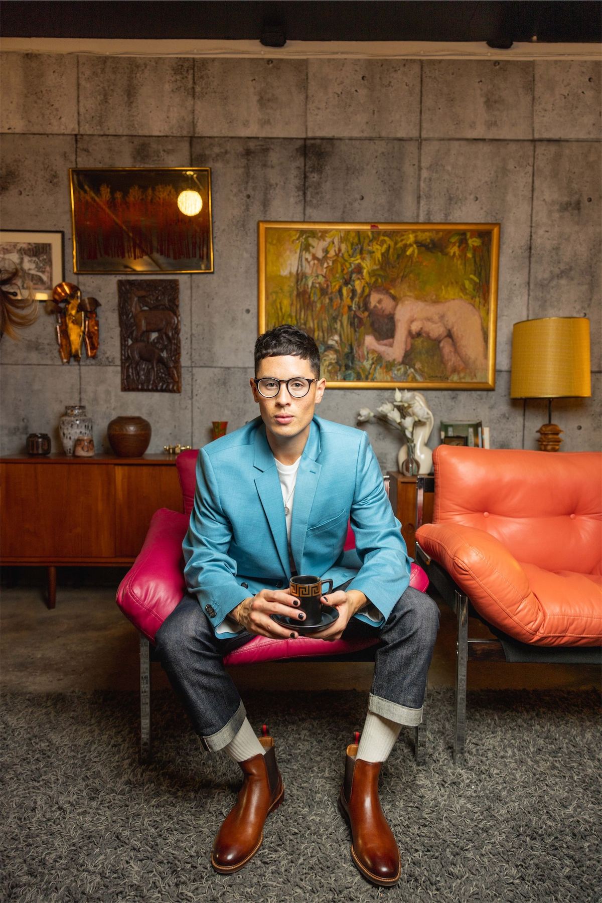 Model in a turquoise blazer and pink shirt seated in a mid-century furnished room
