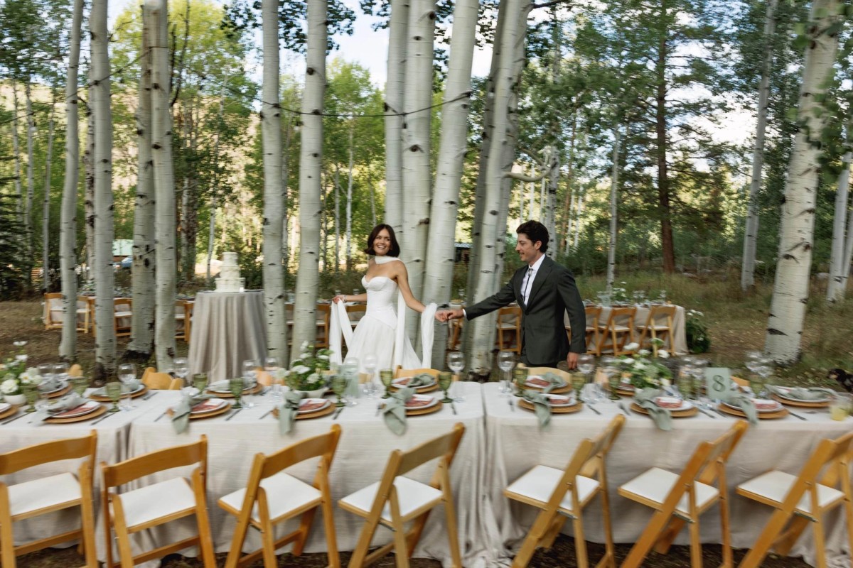 Devin and Owen walk hand in hand past the long dinner table set in the aspens at a mountain ranch among the aspens