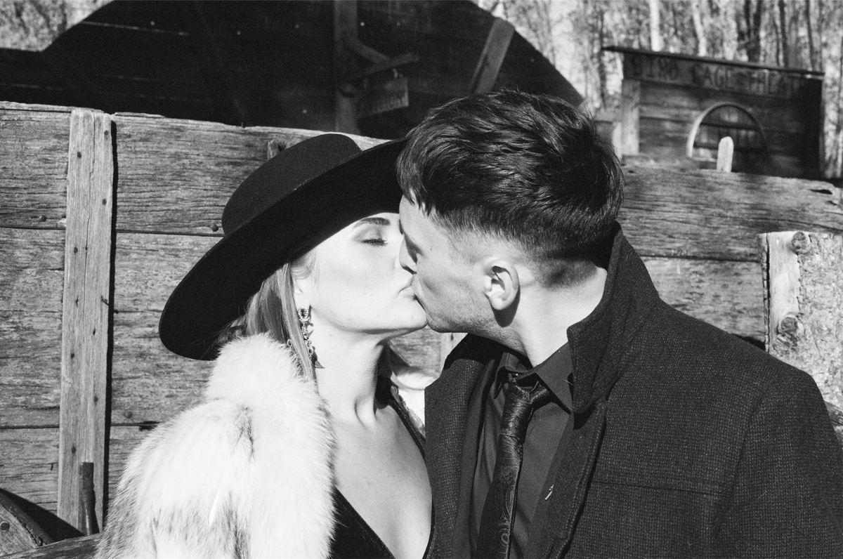 Black and white kiss between the couple in dark coats against a weathered barn wall