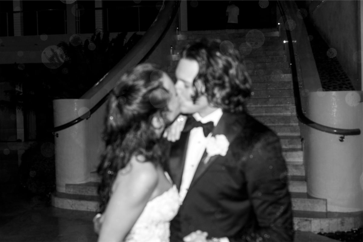 Black and white frame of the couple kissing at the foot of a curved staircase