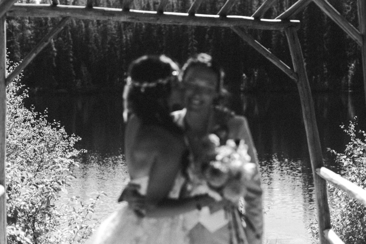 Black and white of the two brides kissing under the timber arch by the lake