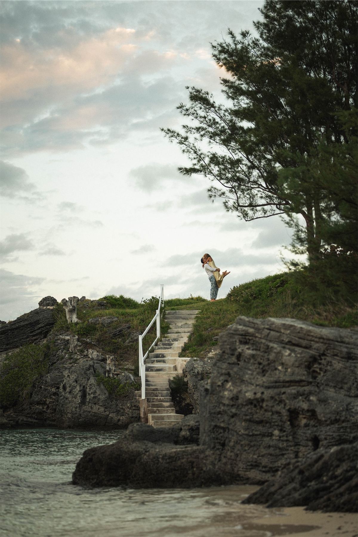 Couple standing small on a rocky bluff above the water at dusk