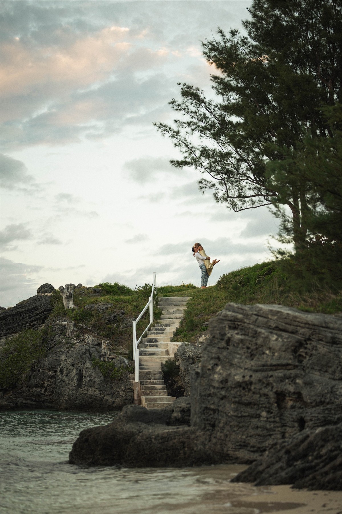 Couple standing small on a rocky bluff above the water at dusk