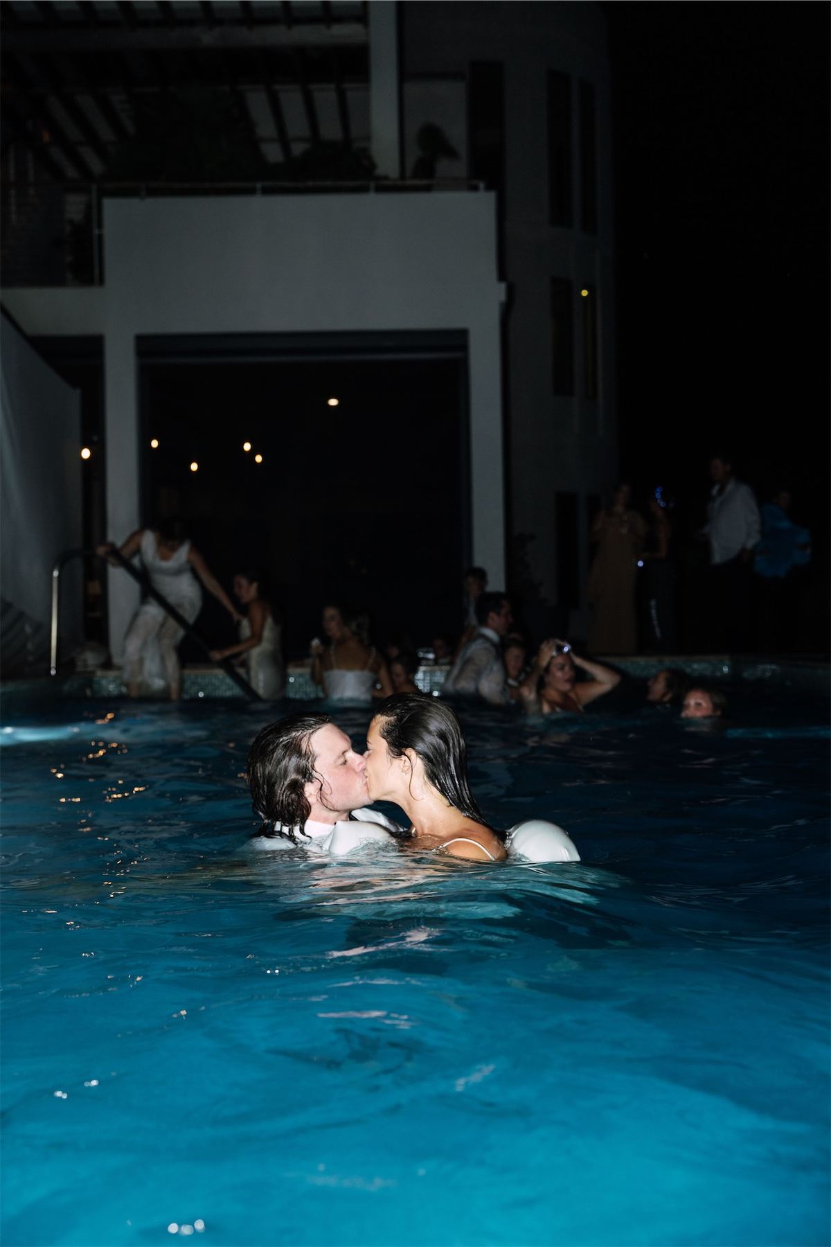 Bride and groom kissing in a lit swimming pool as guests watch from the edge at night