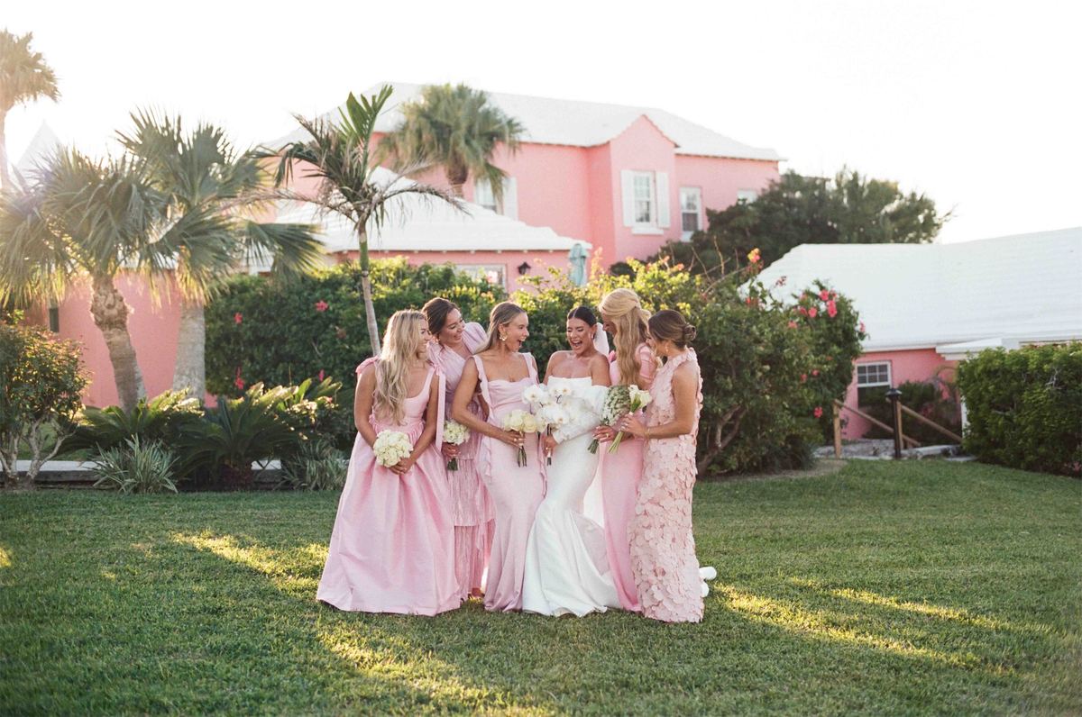 Bridesmaids in pink gathered around the bride beside a coral coloured house