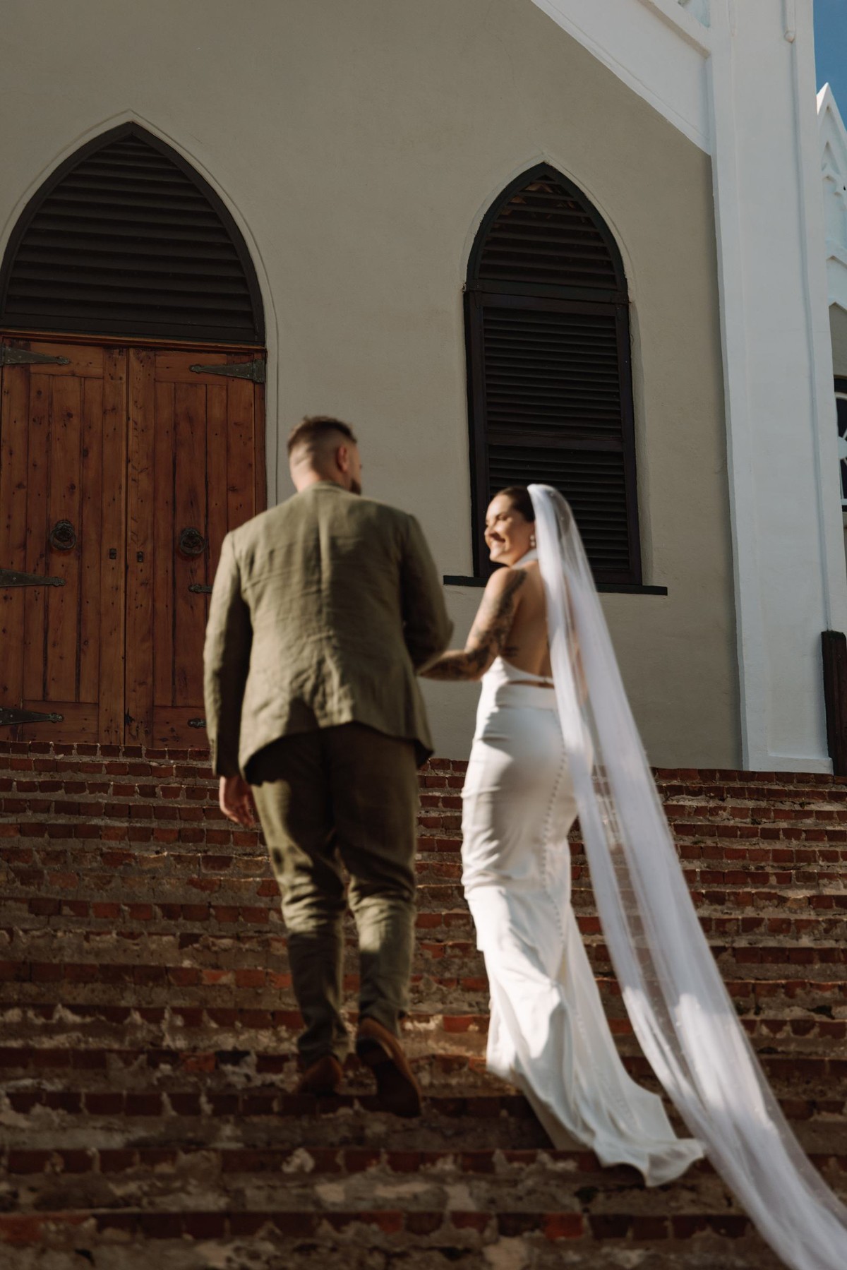 Bride and groom walking up the brick steps to a white Bermuda church, veil trailing behind