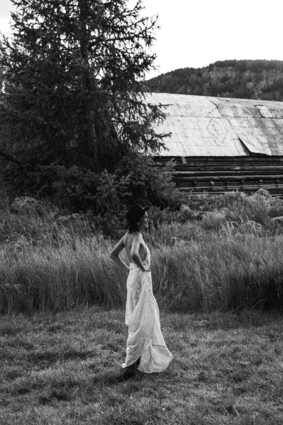 Devin in a white dress and cowboy hat in the long grass by an old barn at a mountain ranch among the aspens, black and white