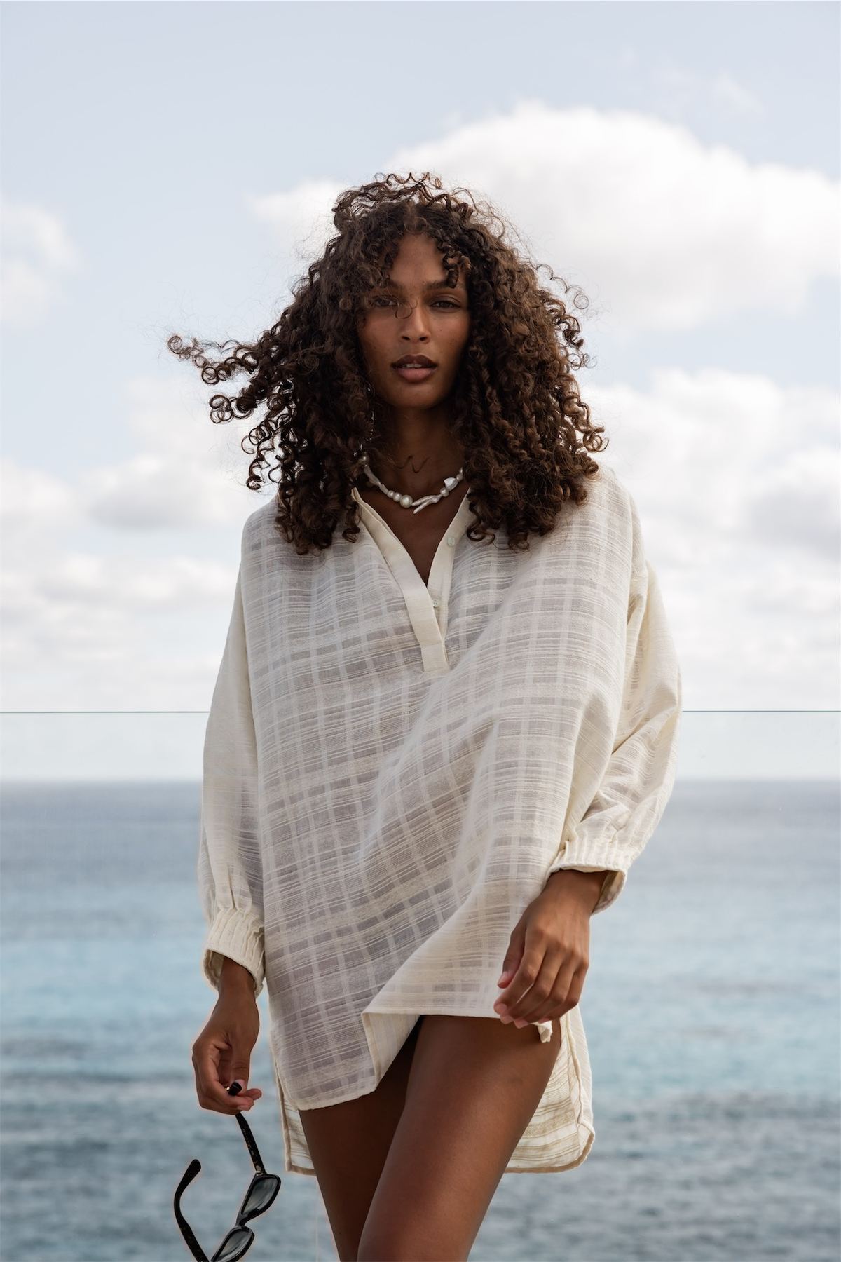 Model in an oversized linen shirt dress holding sunglasses against the open sea